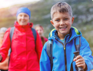Smiling boy in a blue jacket with a backpack leading a group of hikers on an outdoor trail.